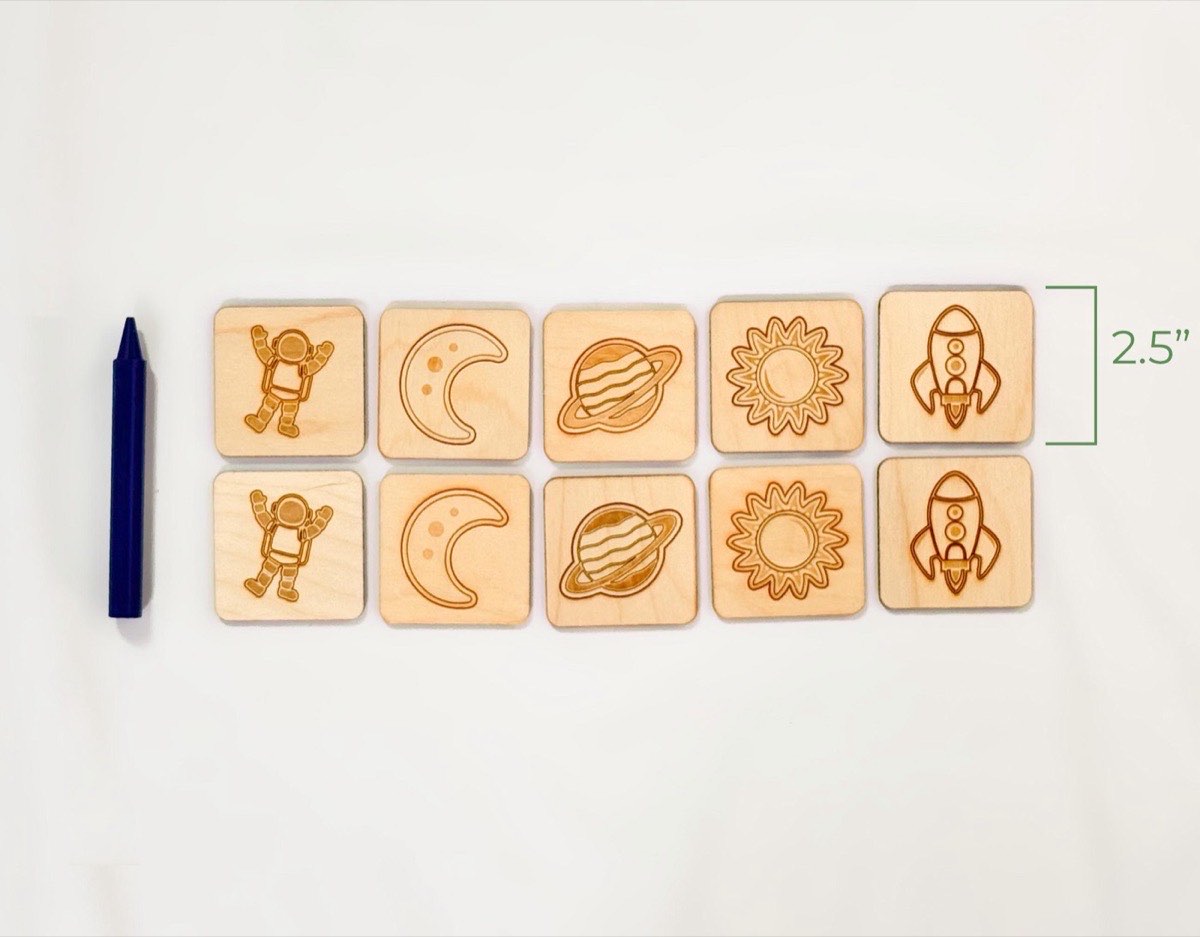 Ten-piece neutral colored wooden space memory match game shown beside jumbo crayon for size reference, featuring astronaut, moon, Saturn, sun, rocket pairs.  On a light white fabric background