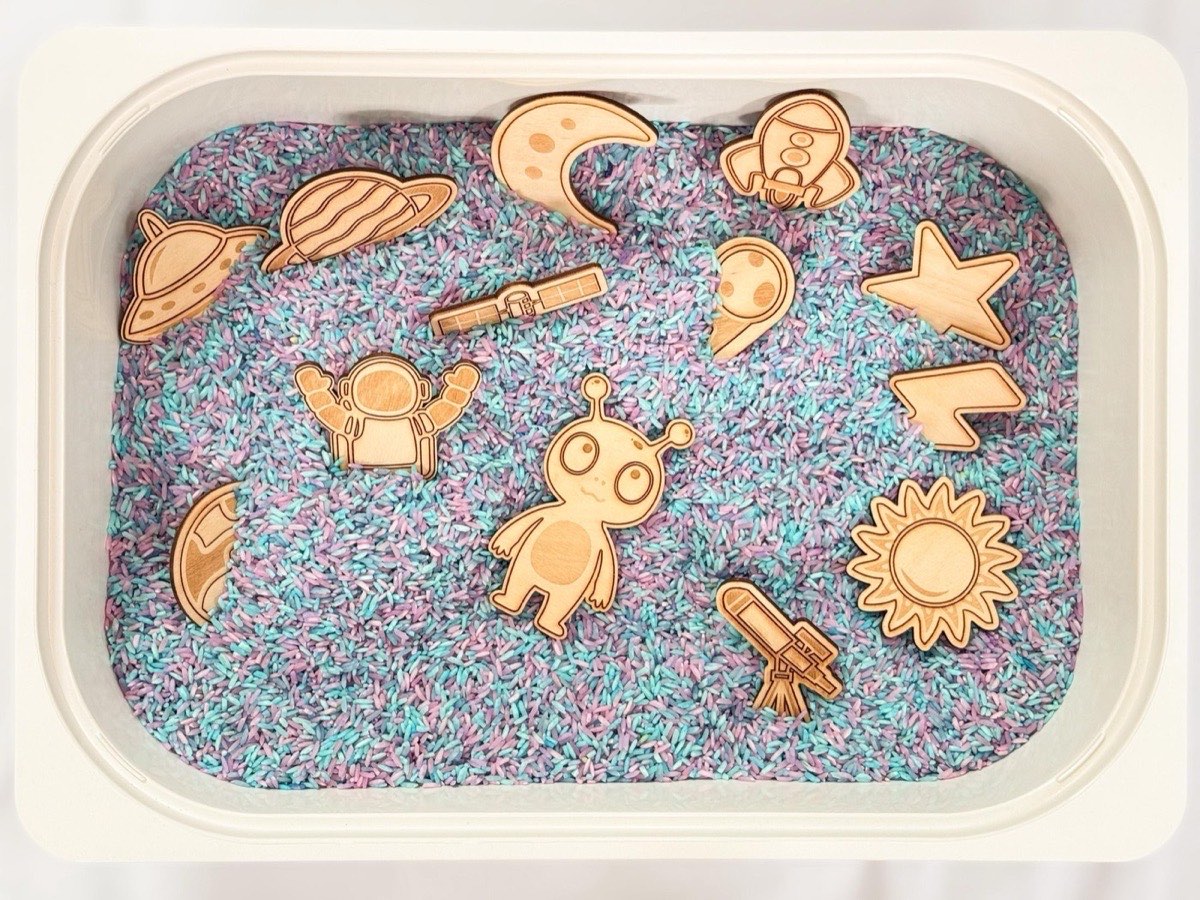 Overhead view of wooden space playscape pieces arranged in a sensory bin  filled with pastel purple, blue, and pink dyed rice. Includes astronaut, alien, rocket, UFO, moon, sun, planets, telescope, satellite, lightning bolt, and star