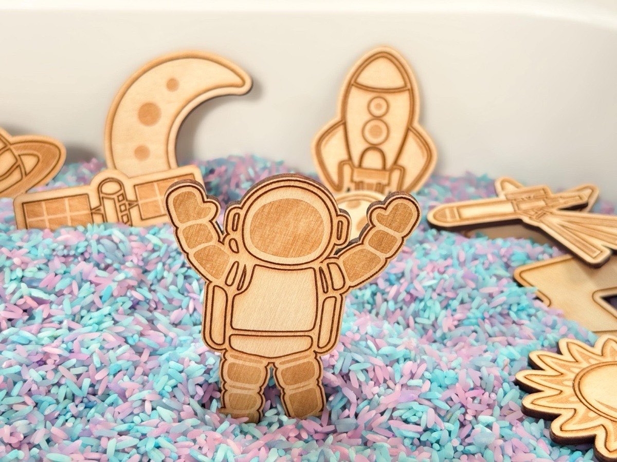 Close-up of laser-engraved wooden astronaut with hands raised standing upright in pastel rainbow sensory rice. Moon, rocket, satellite, telescope, Saturn, and sun pieces visible in the background of a white sensory bin