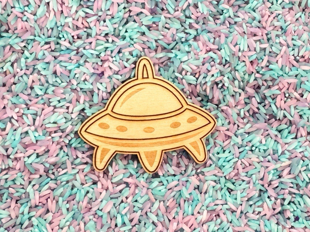 Detailed close-up of a wooden laser-engraved UFO piece resting on pastel pink, teal, and purple dyed sensory rice. Shows fine engraving detail of the flying saucer's dome, portholes, and landing legs on Baltic birch