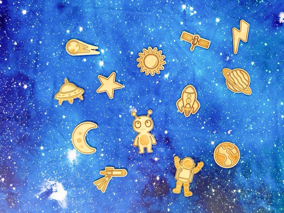 Wooden space-themed Montessori pieces arranged on a starry blue galaxy background. Laser-engraved Baltic birch set includes comet, satellite, lightning bolt, sun, Saturn, UFO, star, rocket, moon, alien, telescope, astronaut, and Earth