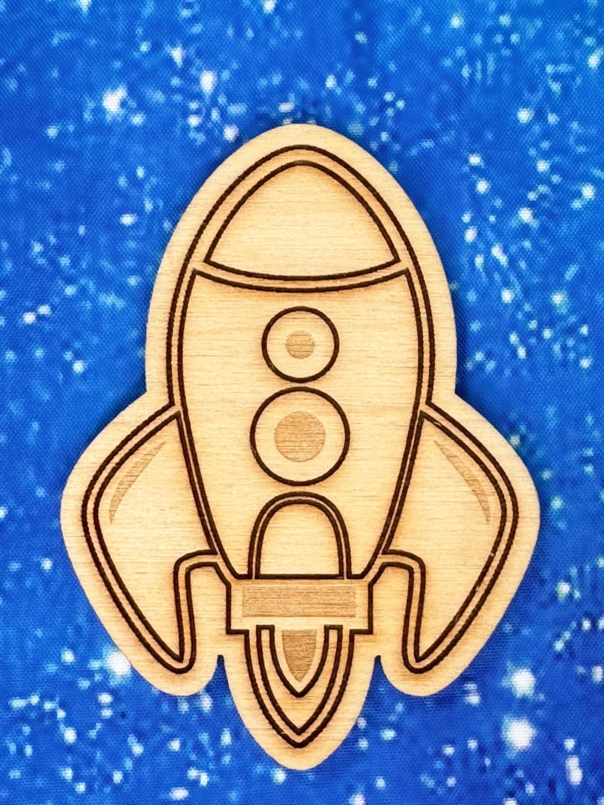 Close-up of a laser-engraved wooden rocket ship on a starry blue galaxy background. Detailed Baltic birch piece featuring engraved windows, fins, and flame exhaust, showcasing fine laser cutting work