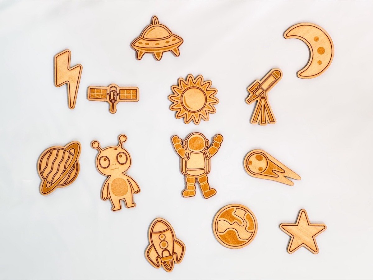 Thirteen-piece wooden space playscape arranged on white background. Laser-engraved Baltic birch pieces include UFO, moon, lightning bolt, satellite, sun, telescope, Saturn, alien, astronaut, comet, rocket, Earth, and star. Top-down flat lay on white fabric background