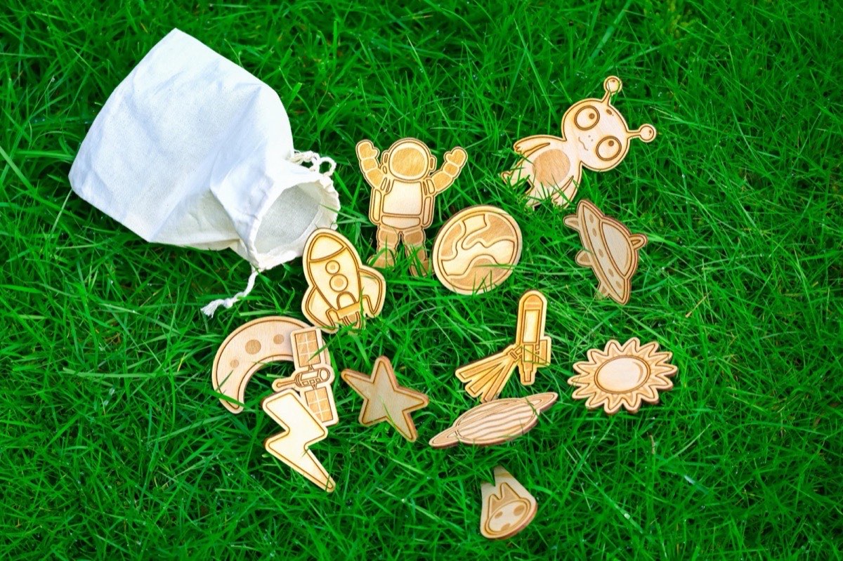 Wooden space-themed Montessori toys spilling from a natural cotton drawstring bag onto bright green grass. Set includes astronaut, alien, rocket, planet, UFO, moon, star, sun, lightning bolt, satellite, and comet, all laser-engraved on Baltic birch