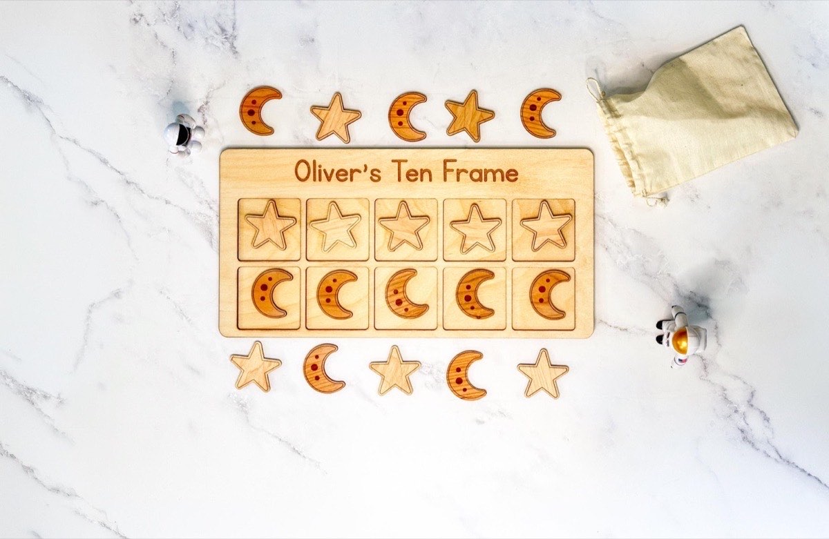 Personalized space ten frame engraved 'Oliver's Ten Frame' on white marble, five star counters in the top row and five crescent moon counters below, extra pieces, astronaut figurines, and a muslin drawstring bag around the board