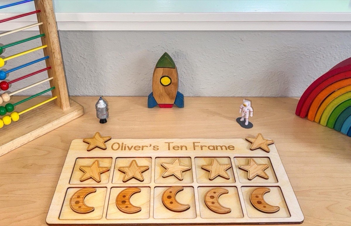 Space ten frame on a playroom shelf beneath a wooden rocket ship, astronaut and robot figurines, rainbow stacker, and abacus, ten star and moon counters filling the personalized board