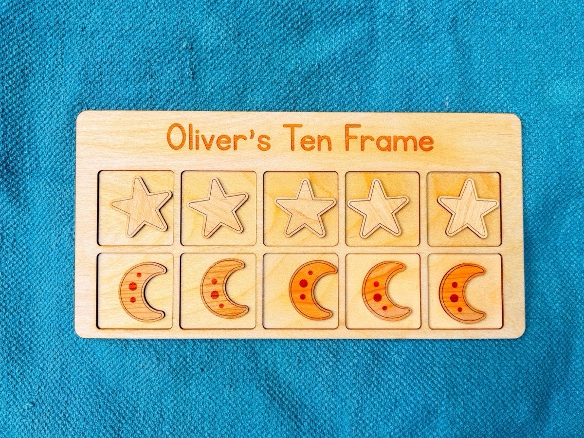 Overhead view of a space-themed ten frame on a teal mat, a row of five star counters above a row of five moon counters, board personalized with a child's name in engraved lettering