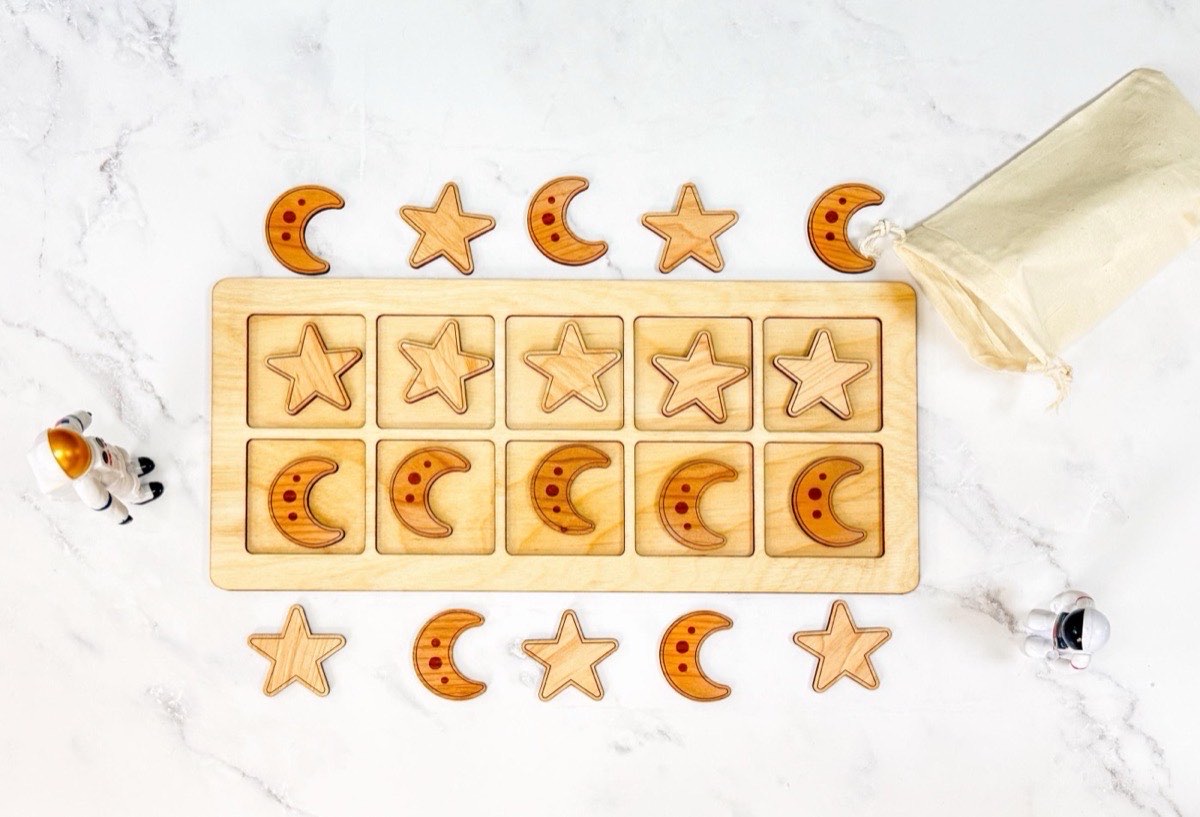 Non-personalized wooden ten frame with five laser-engraved stars and five spotted crescent moons in two rows of neutral Baltic birch squares, surrounded by extra space counters, toy astronauts, and a natural muslin bag