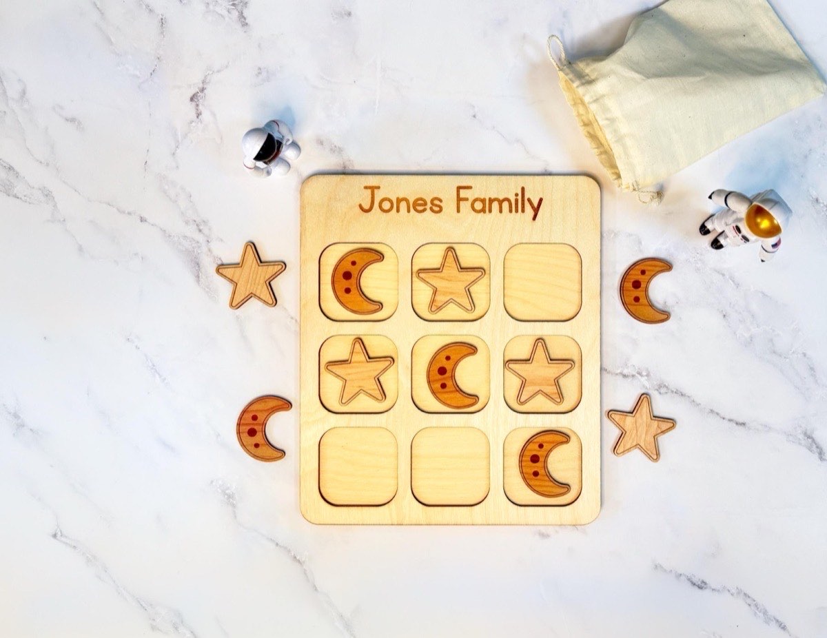 Personalized space tic tac toe board engraved 'Jones Family' on marble, laser-engraved crescent moon and star pieces mid-game with astronaut figurines and a muslin drawstring bag nearby