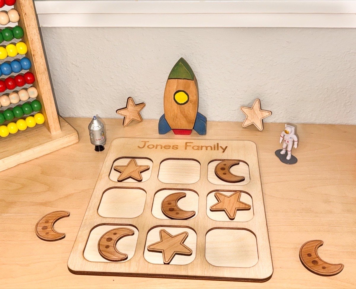 Space tic tac toe on a playroom desk with a wooden rocket ship, astronaut and robot figurines, and abacus, moon and star pieces mid-game on the family-name board