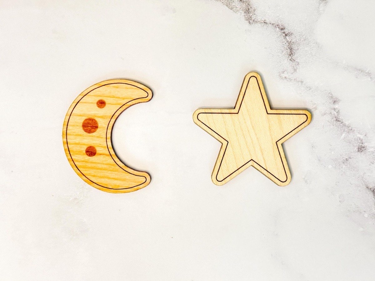Close-up of two wooden tic tac toe pieces: a crescent moon with engraved crater dots in warmer wood and a five-pointed star in light neutral wood