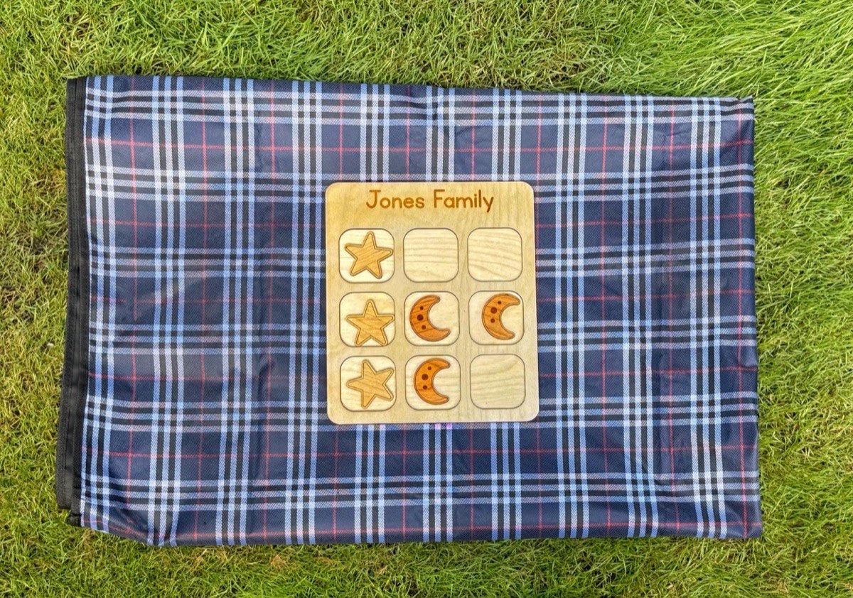 Space tic tac toe board on a navy plaid picnic blanket in grass, star and moon playing pieces spread across the 'Jones Family' grid for an outdoor game