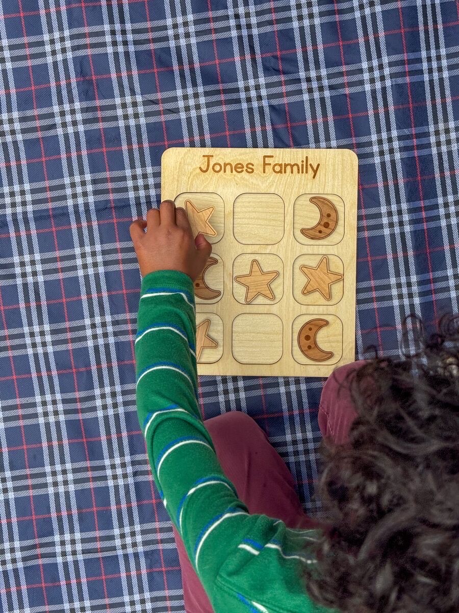 Overhead view of a child's hand placing a maple star on a tic tac toe board engraved 'Jones Family' on a navy plaid blanket, four stars and three cherry crescent moons with engraved craters filling the three by three grid